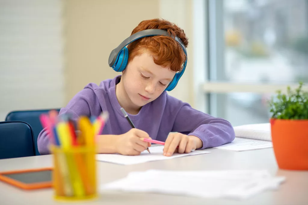 Boy wearing headphones is sitting and doing some work at a desk. 