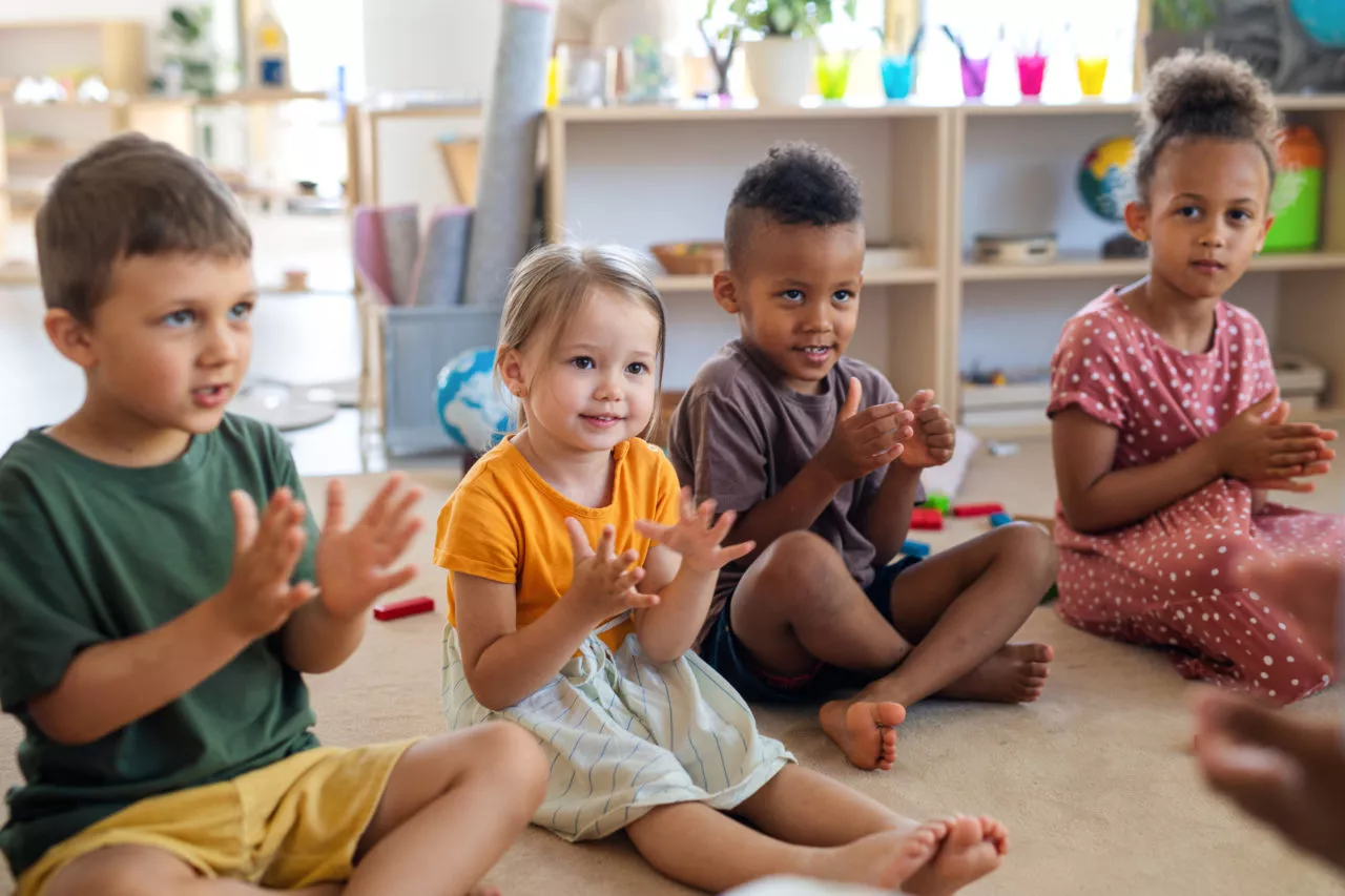 Early literacy in preschool: A group of small nursery school children sitting on floor indoors in classroom, clapping to syllables