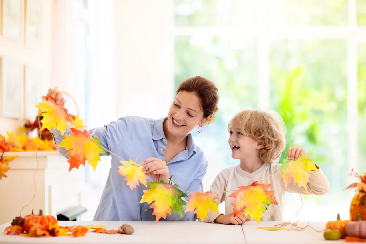 Autumn home decoration. Family decorating house with colorful maple leaves banner. Thanksgiving or Halloween celebration in white living room. Child with fall leaf. Creative crafts ideas with fall leaves