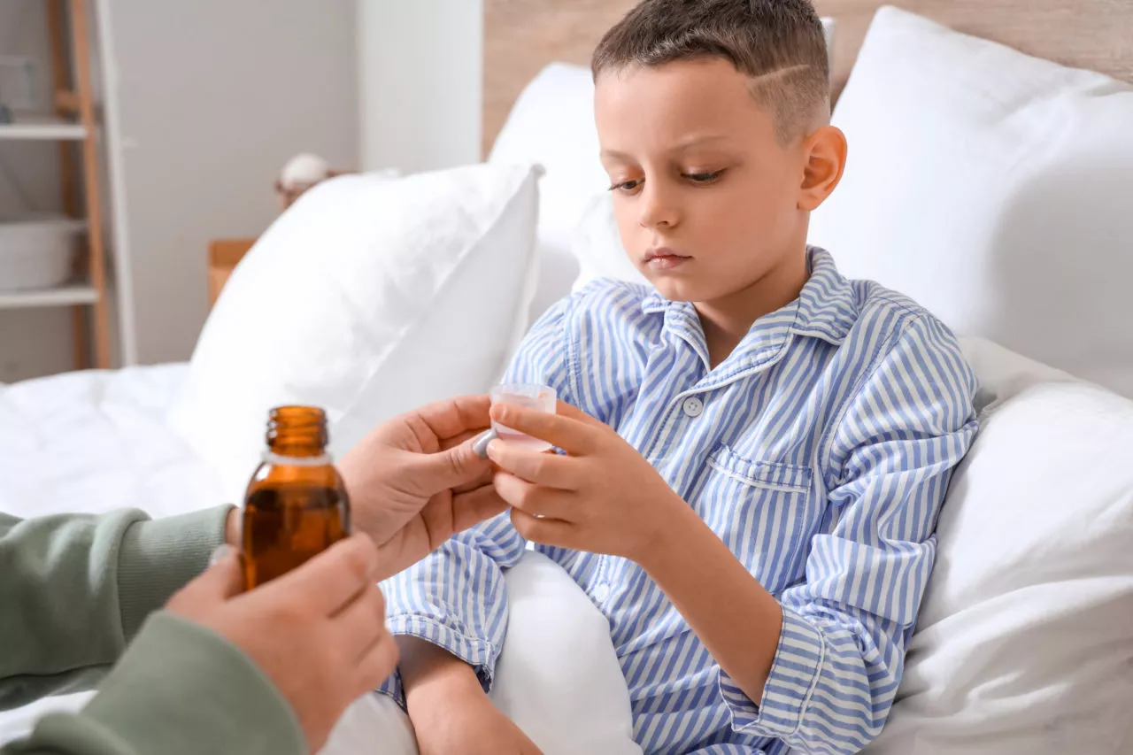 when should kids stay home from school: Ill little boy taking cough syrup from his mother in bedroom
