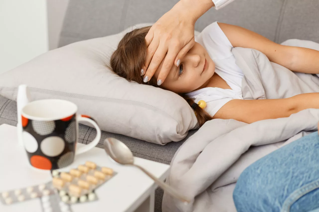 when should kids stay home from school: Indoor shot of little unhealthy girl lying on cough, mother taking care of her sick daughter, lovely mommy touching kid's forehead to measure temperature.