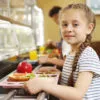Nutrition affects learning in children: Little girl with tray of tasty food in school canteen, selective focus. Healthy meal with fruits, vegetables, and grains supporting children’s learning and focus