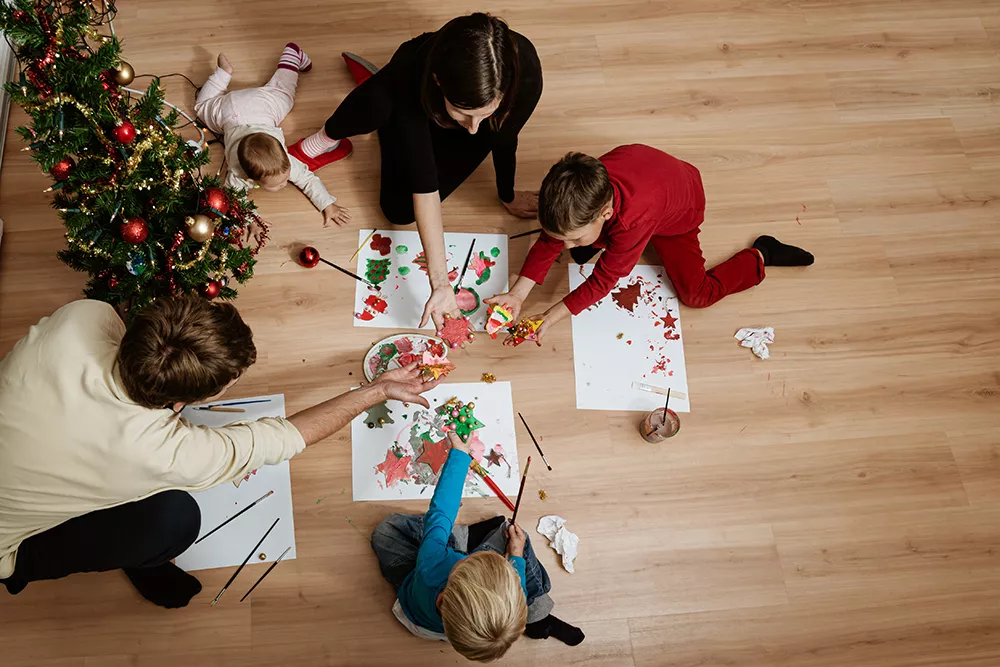 Top view of family of five sitting on living room floor next to a christmas tree making holiday ornaments and decoration together.