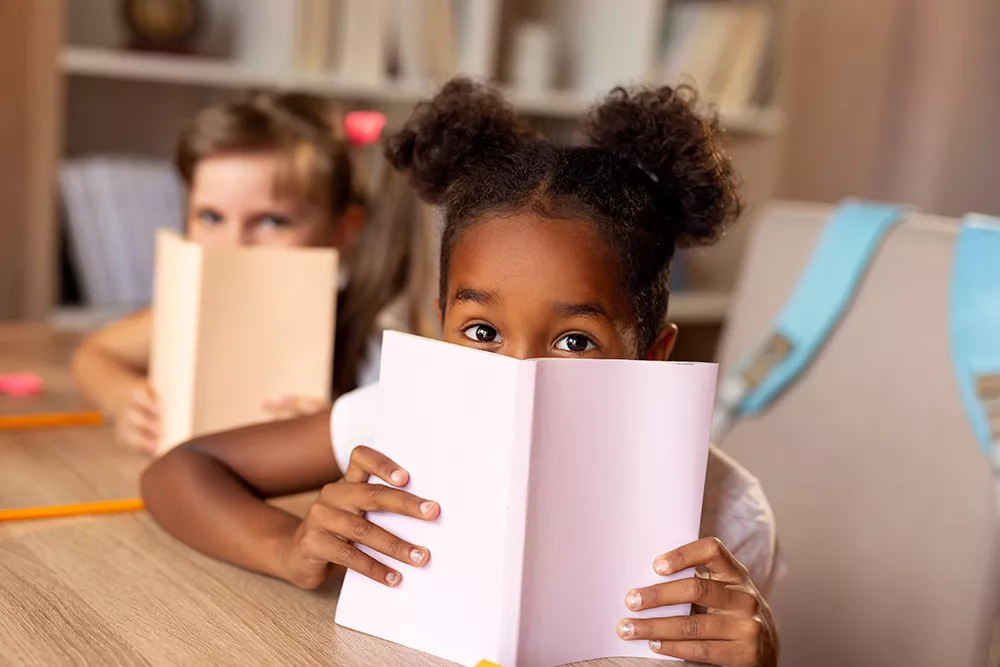 Two little girls sitting at a desk, hiding behind books and peeking