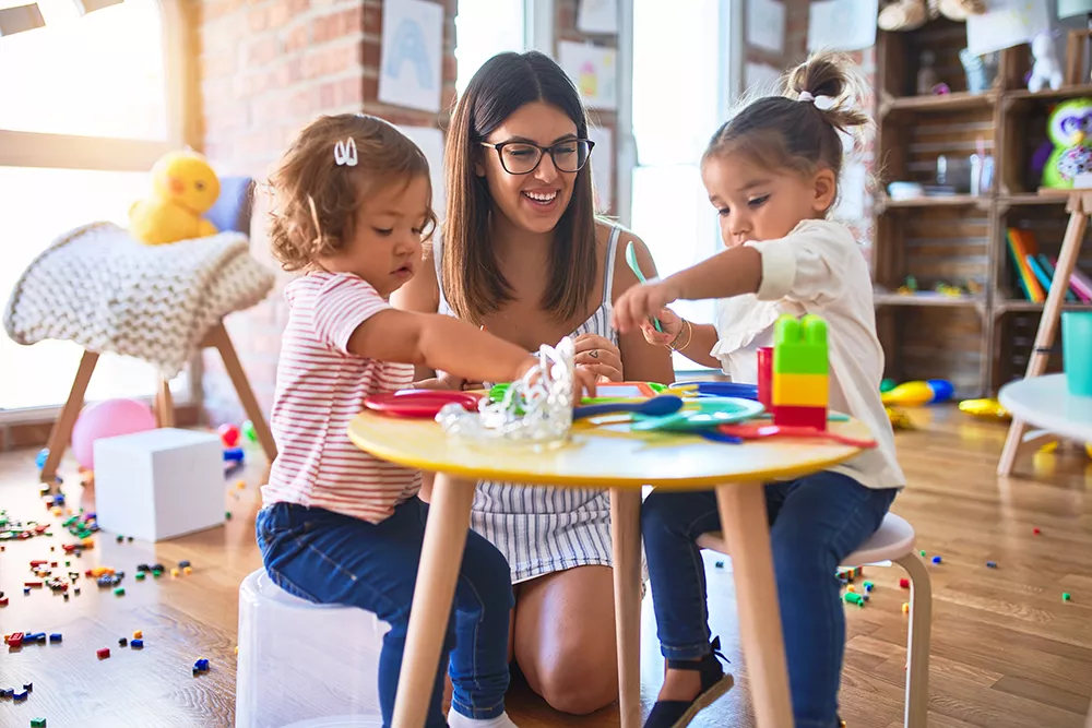 Teacher and toddlers playing meals using plastic food and cutlery toy at daycare.