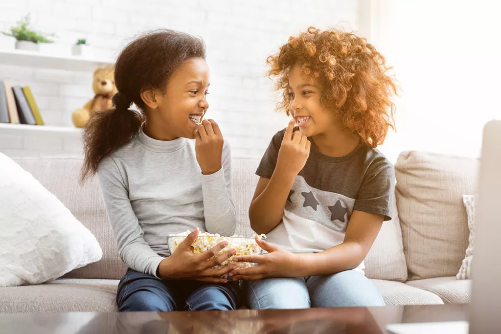 Kids eating popcorn, looking at each other, sitting on sofa.