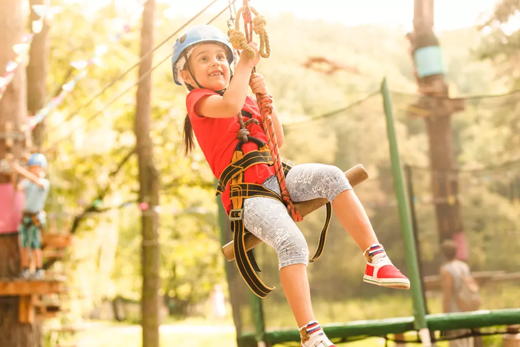 Brave young girl in helmet climbs on tree tops in amusement rope park.