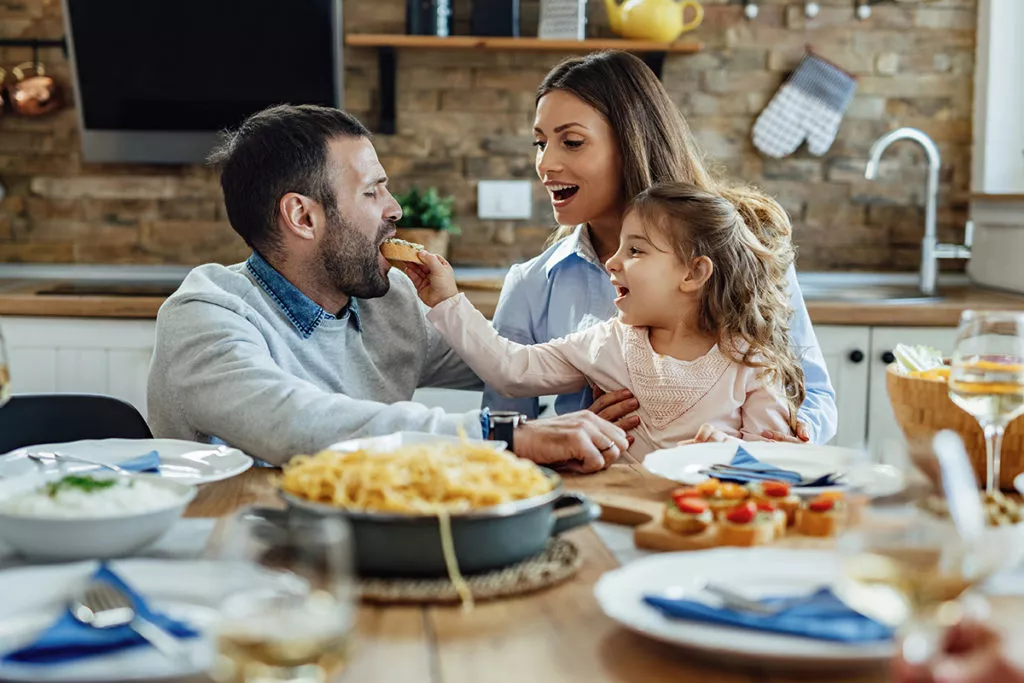 Happy family having fun during lunch at dining table. Little girl is feeding her father.