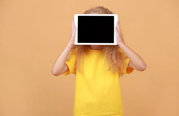 Kid hiding behind digital tablet with empty screen over yellow studio background.