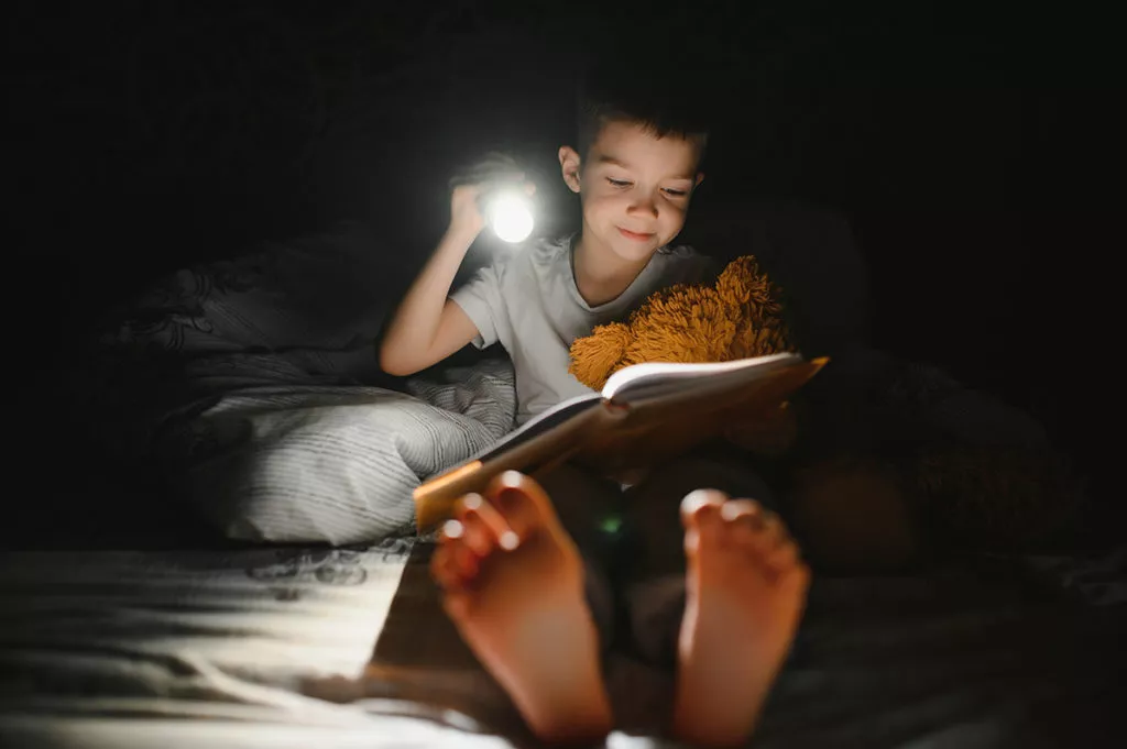 Child reading book in bed in the dark with a flashlight.