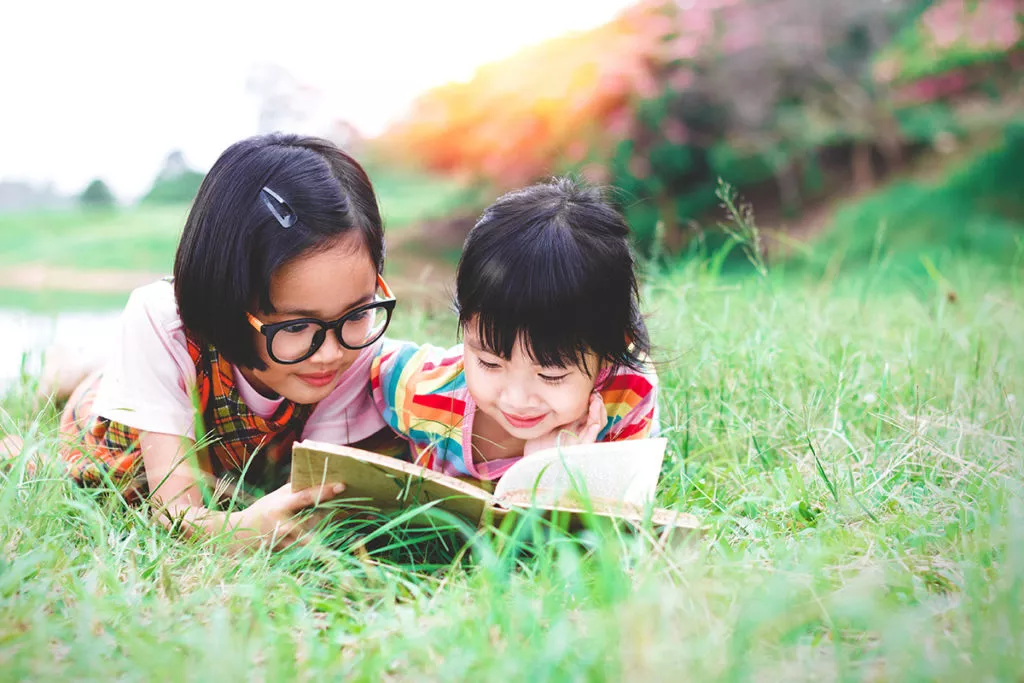Two little girls with glasses lying on the grass reading a book in garden at summer time