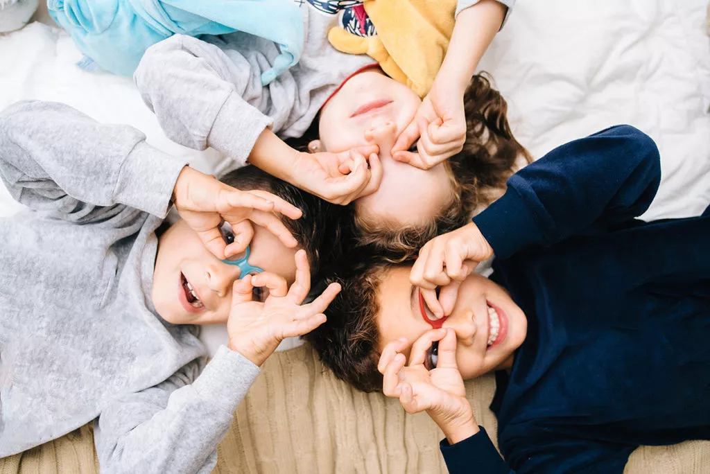 Portrait of three children smiling and making glasses with their fingers lying on the floor.