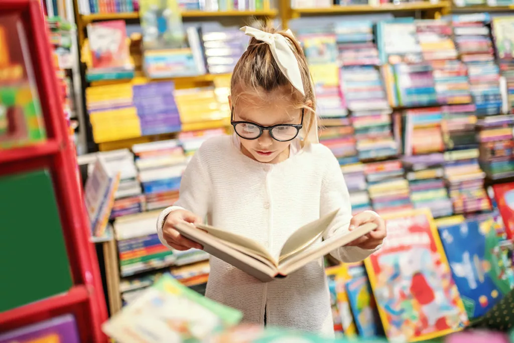 Adorable little girl with eyeglasses standing in bookstore and reading interesting book. All around are books on shelves.