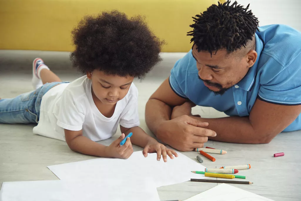 father and child laying on the floor and coloring together.
