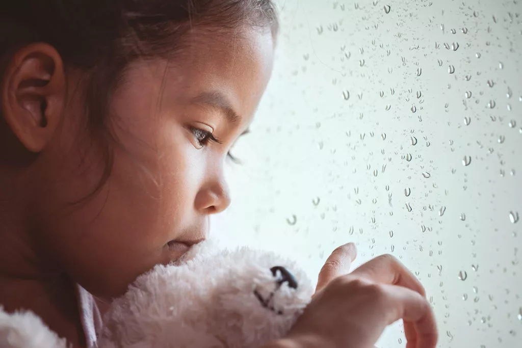 Sad young girl with her teddy looking outside while she is stuck indoors because of bad weather.