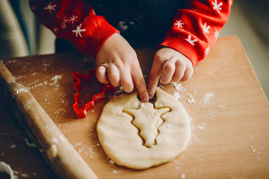 From above shot of kid cutting tree-shaped biscuit from soft dough while preparing pastry for Christmas in the kitchen during his holiday break from school.