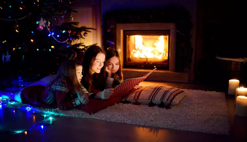 Happy young mother and her daughters reading a story book together by a fireplace in a cozy dark living room during Christmas break.