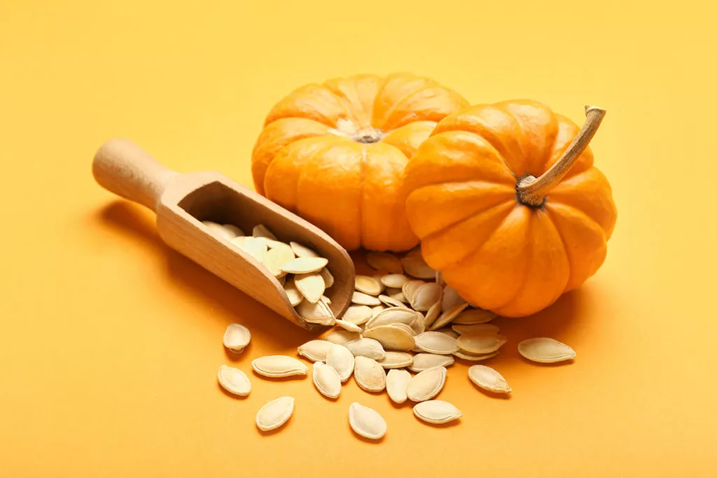 Pumpkin seeds with a scoop and a couple of small pumpkins on a yellow backgound.