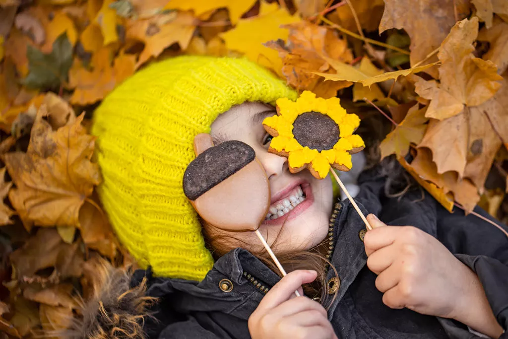 A little funny girl in a yellow hat lies in the autumn foliage and holds gingerbread in her hands.