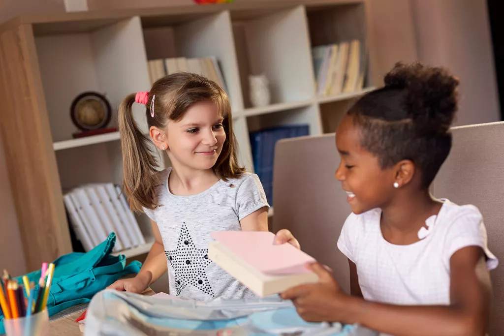 Two girls at school happy and socializing while sharing some books at a table.