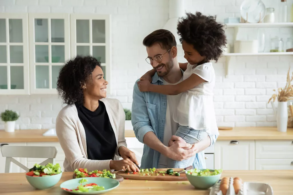 A family in the kitchen, the dad is holding the child and the mom is cooking dinner and hapilly looking up at them.
