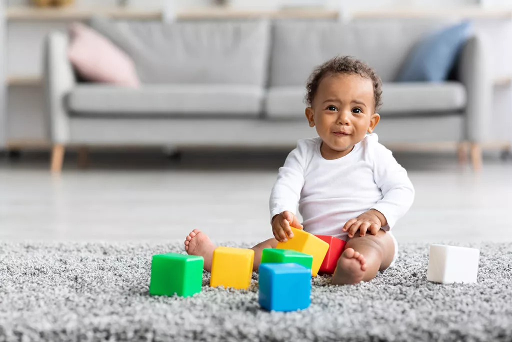 Little baby sitting on the rug and playing with blocks in front of a couch in the living room.