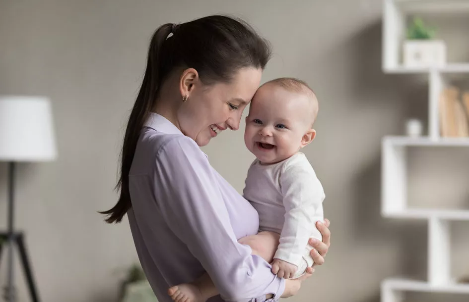 Young mom and cheerful newborn in bodysuit laughing together.