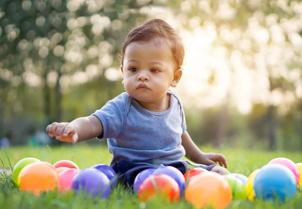 Cute baby playing with colorful balls in green grass while at an infant daycare in Rockville, MD.