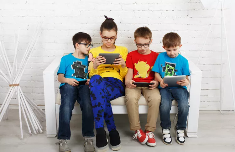 group of kids of various ages sitting on a bench and looking at different tablets or screens.