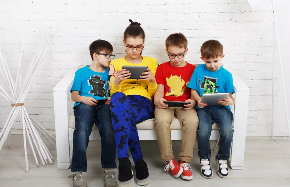 group of kids of various ages sitting on a bench and looking at different tablets or screens.