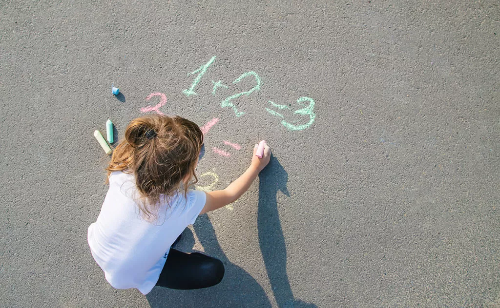 Preschool girl writing simple math problems in chalk on the ground as an Engaging Activity to Teach Early Numeracy Skills