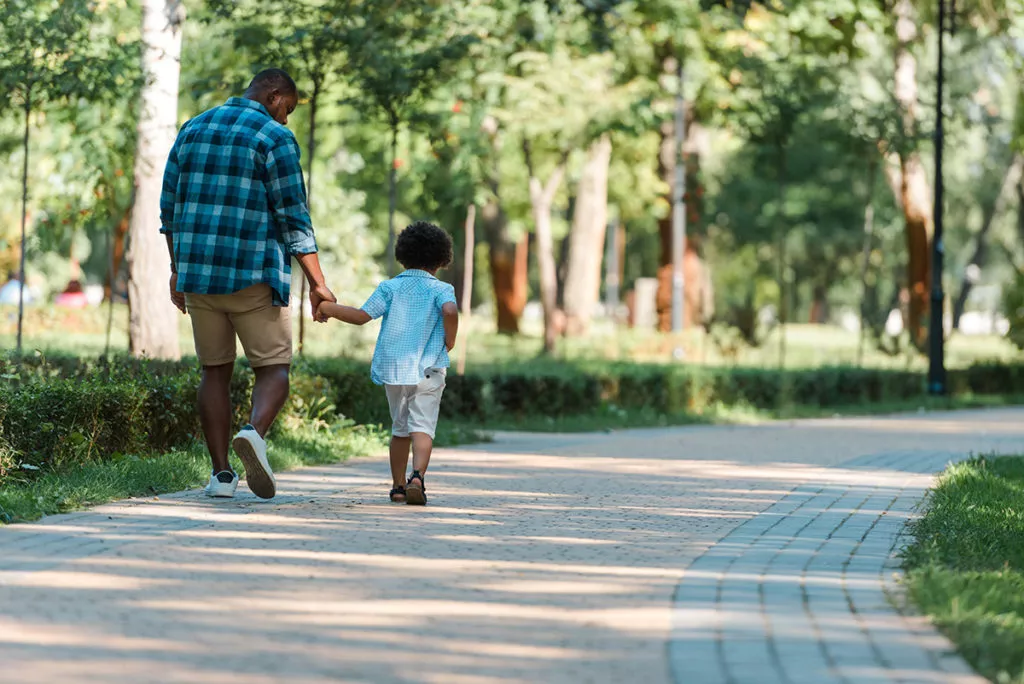 Back view of preschool boy holding hands with his father and walking in a park.