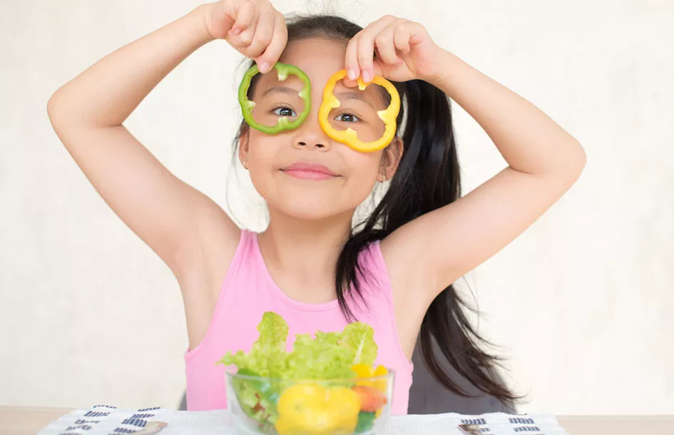 Young girl holding yellow pepper circles over her eyes and showing the importance of well balanced nutrition for kids with busy parents.
