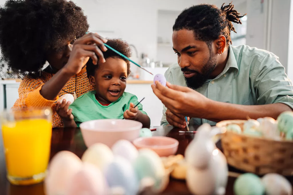 Happy parents with toddler son painting colorful eggs together on a rainy spring day.