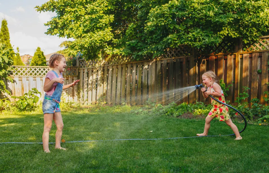 Two girls splashing each other with gardening house on backyard on summer day. Children playing with water outside at sunset. Candid moment, lifestyle activity. Life friendship of sisters siblings.