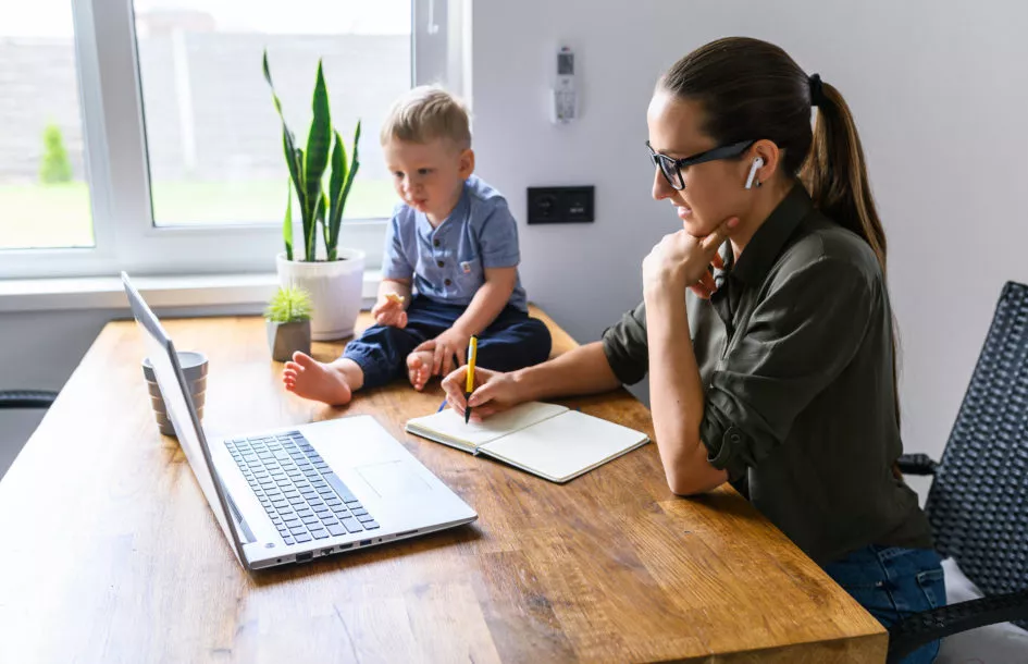 Young mother watching online webinars, online classes with a child sitting near. Mom studying online and writing in notebook