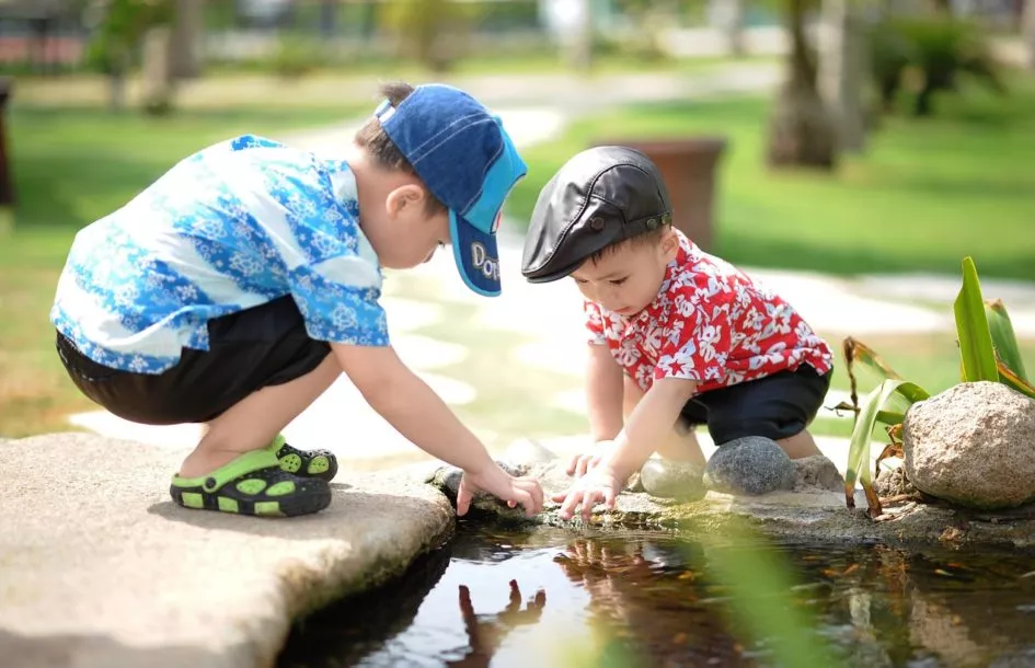 Two young boys are crouched near the edge of a small pond, reaching into the water with their hands. One boy is wearing a blue patterned shirt, black shorts, a blue cap, and green sandals. The other boy is dressed in a red floral shirt, black shorts, and a black leather cap. They are outdoors in a park-like setting with grass, rocks, and plants around them.