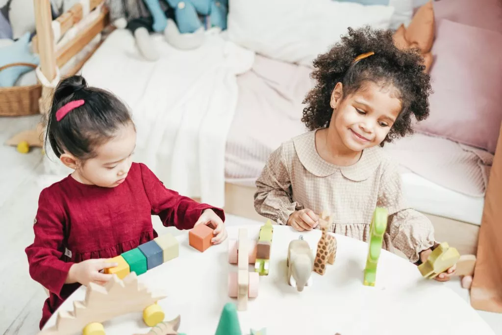 Two young girls are sitting at a small round table indoors, playing with wooden toys and blocks. One girl, wearing a red dress, is focused on stacking colorful blocks. The other girl, dressed in a beige checkered dress with a Peter Pan collar, is smiling while holding a green dinosaur toy. The room has soft furnishings, including a bed with pillows and stuffed animals in the background.