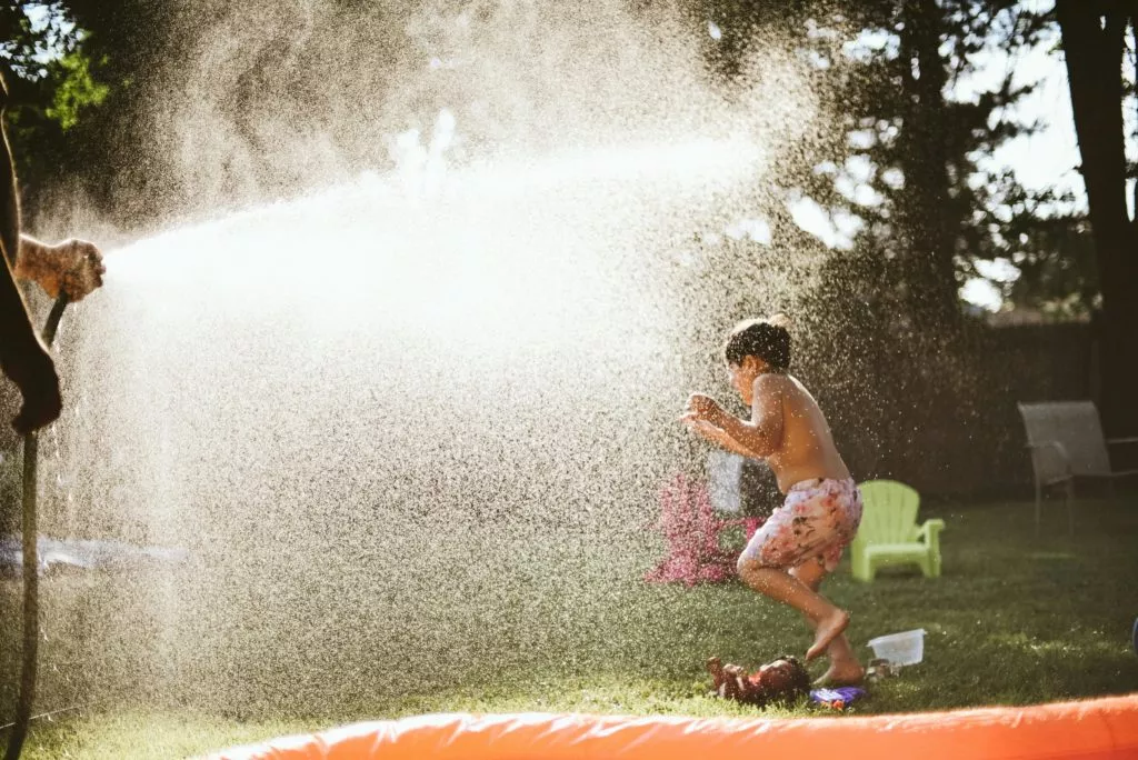 A young person, appearing to be a child, is joyfully playing in the spray of a water hose held by an adult, in a sunny backyard with an inflatable pool visible in the foreground.