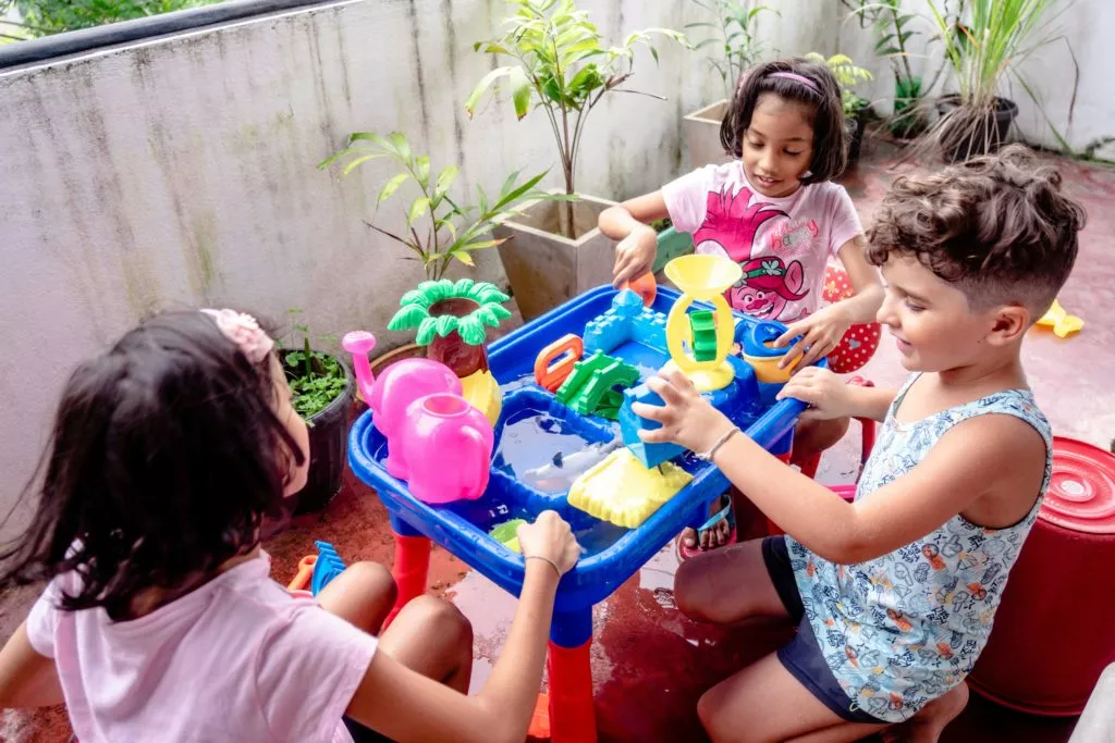 Three children are gathered around a blue water table filled with water and various colorful plastic toys, engaged in play on what appears to be a patio or balcony with potted plants. The children seem to be enjoying a water activity together.
