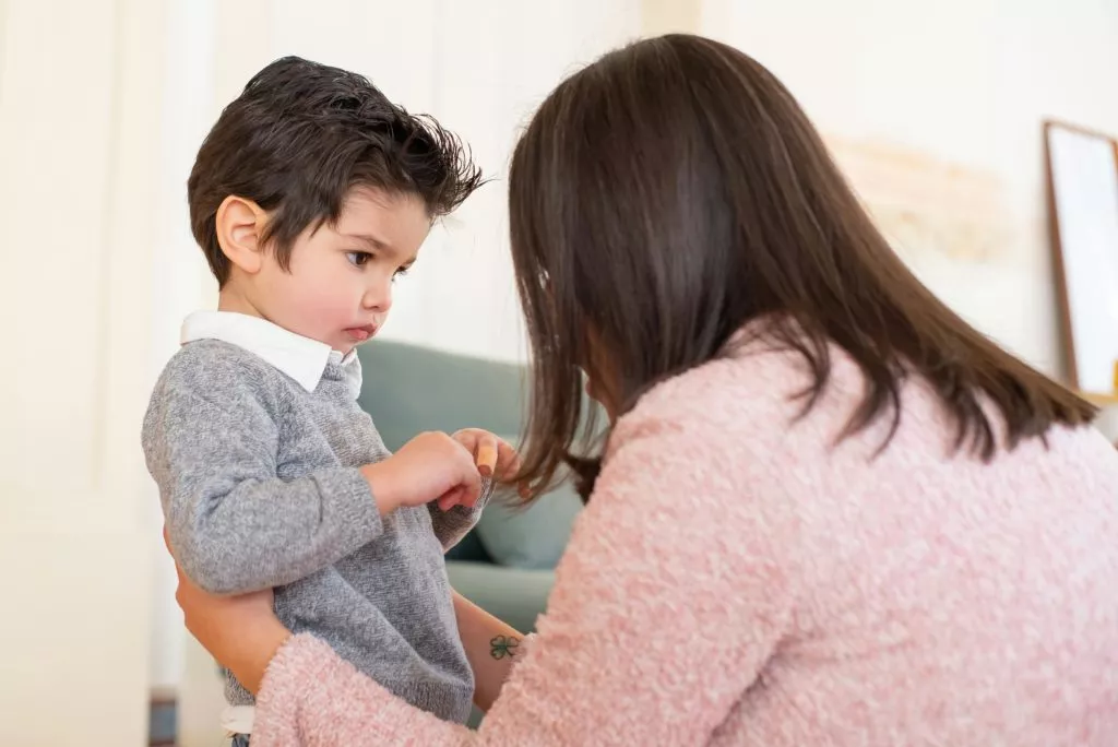 A young child with dark hair and a grey sweater looks attentively at an adult with long brown hair and a pink sweater, who is partially visible from the back.