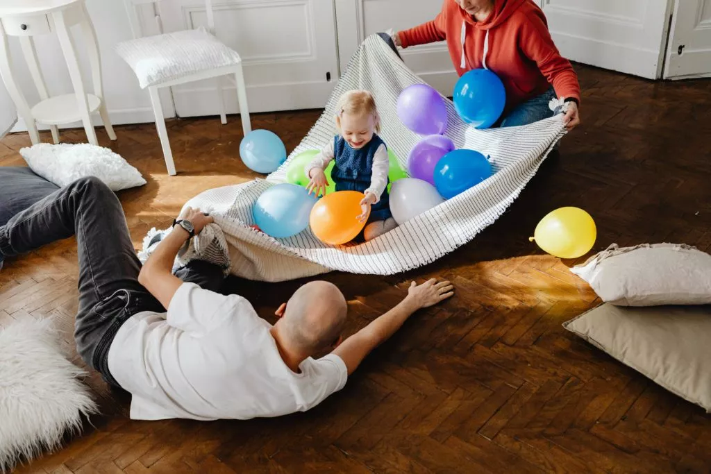 A family of three, including two adults and a baby, is playing with colorful balloons on a blanket in a bright living room.