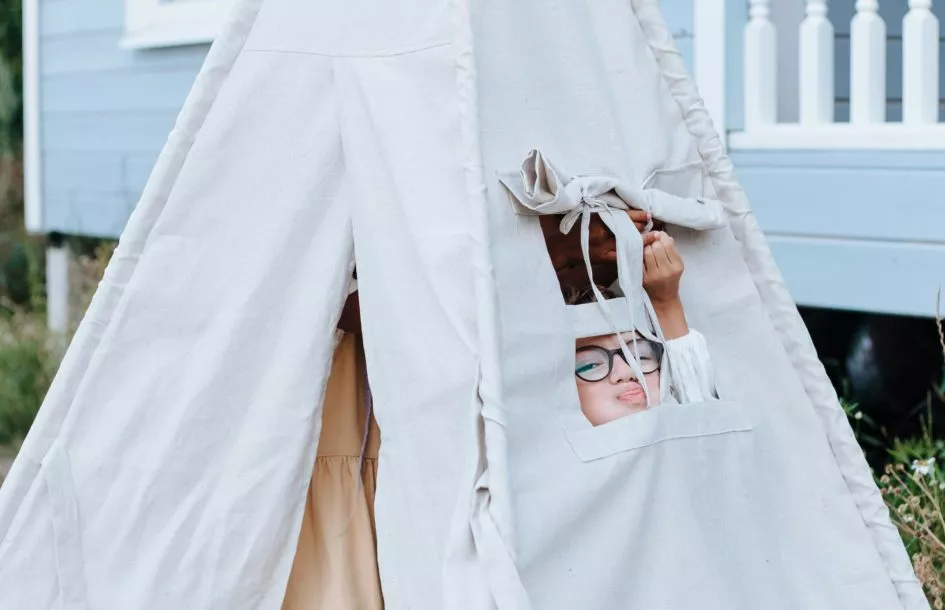 A child wearing glasses peeks out from the small window of a light-colored teepee tent set up outdoors.