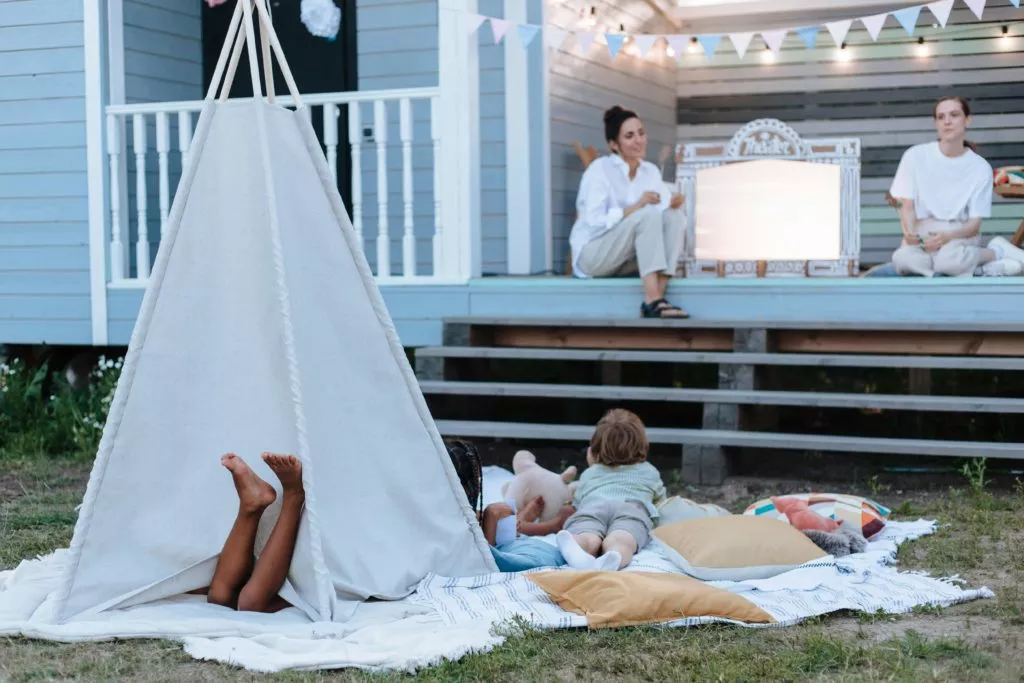 A light teepee on a blanket holds a child with darker skin tone, bare feet peeking out. Another child looks on as two adults sit on the porch of a blue house, decorated with festive lights and flags, observing the scene. Pillows and toys add to the cozy atmosphere.