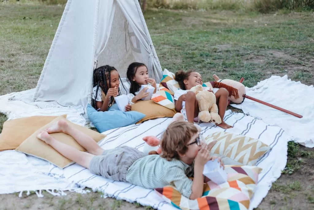 Four children, appearing to be diverse, relax on a blanket with pillows outside a teepee, seemingly enjoying snacks and each other's company.