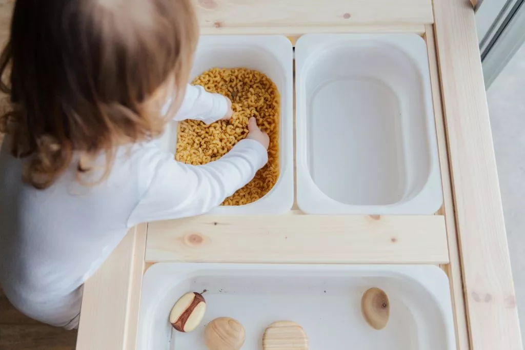 An overhead view shows a child with light brown hair reaching into a white container of yellow pasta. The container sits on a light wooden sensory table. Other containers hold wooden sensory toys.