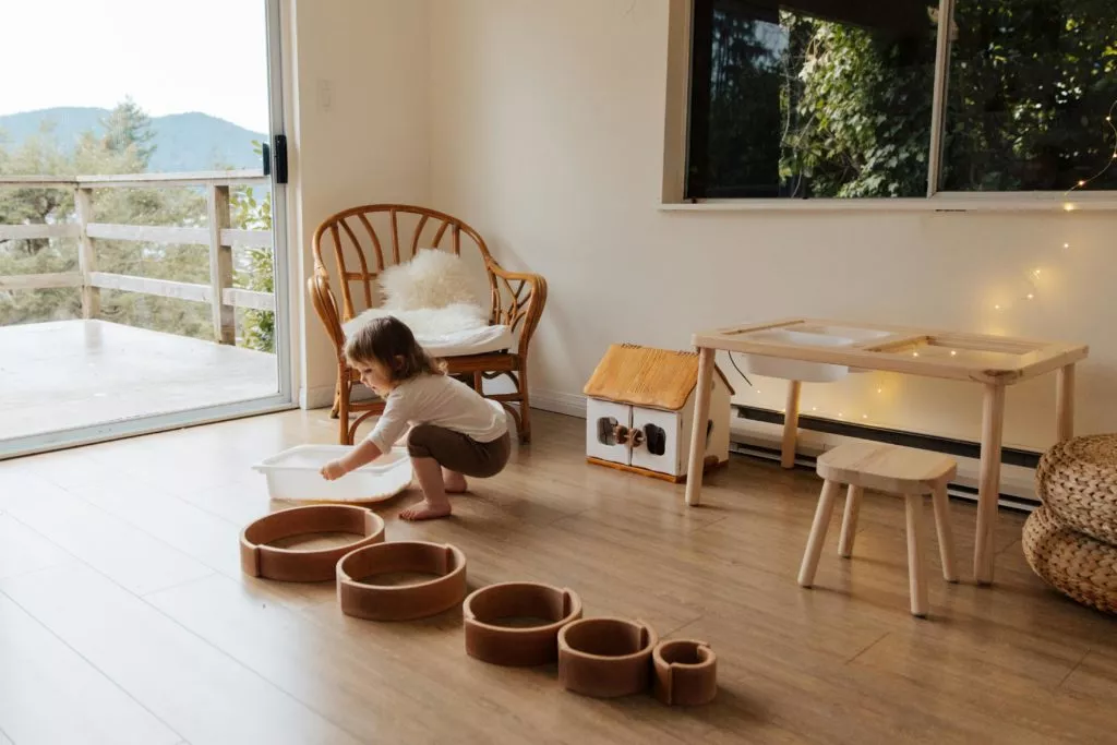 A young child plays with water and wooden rings on the floor of a sunlit room. The room includes a wooden chair, playhouse, light table, and a balcony door with a scenic view. String lights decorate the window.