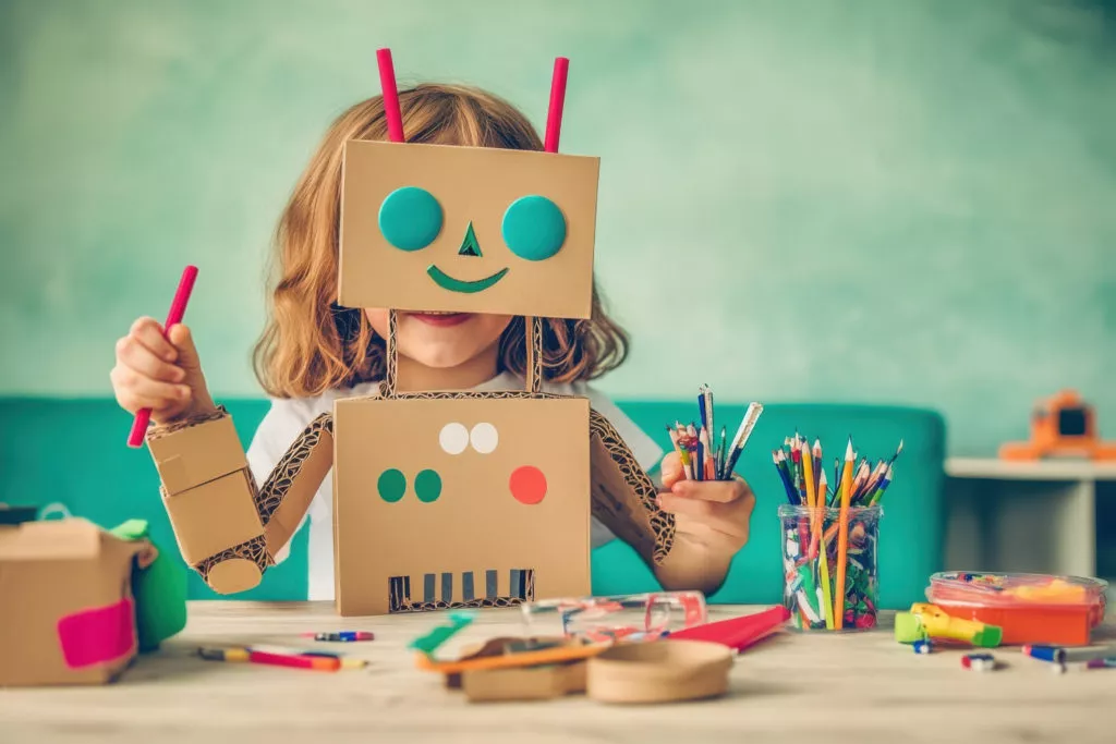 A child joyfully constructs a playful robot from cardboard and art supplies at a crafting table.