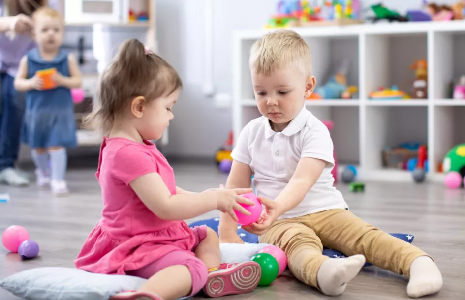Supporting toddler social skills: a girl and boy toddler pictured sharing a ball the girl is giving to him in the playroom