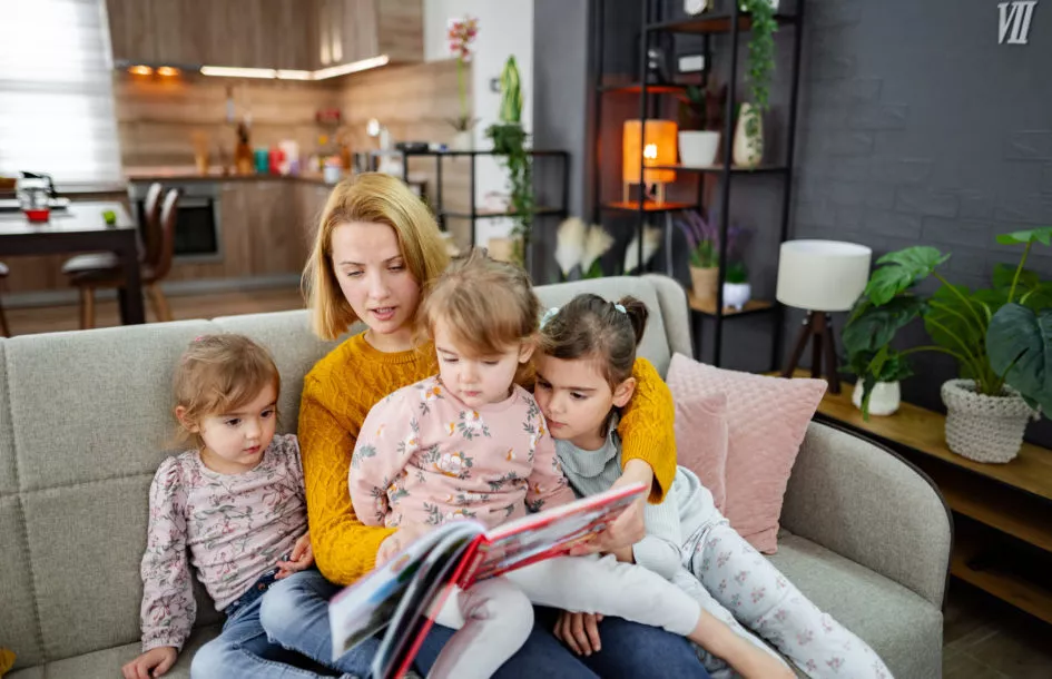 A mother and her three daughters are sitting on the couch in the living room, listening carefully to their mother reading a story, story telling activities for preschoolers